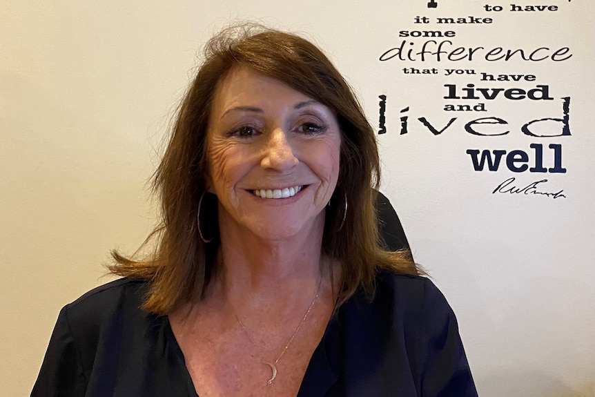 A woman smiles at the camera and there are words on the wall behind her offering hopeful words