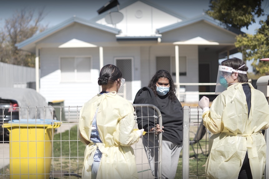 Two health workers in PPE talk to a woman over a fence in her front yard