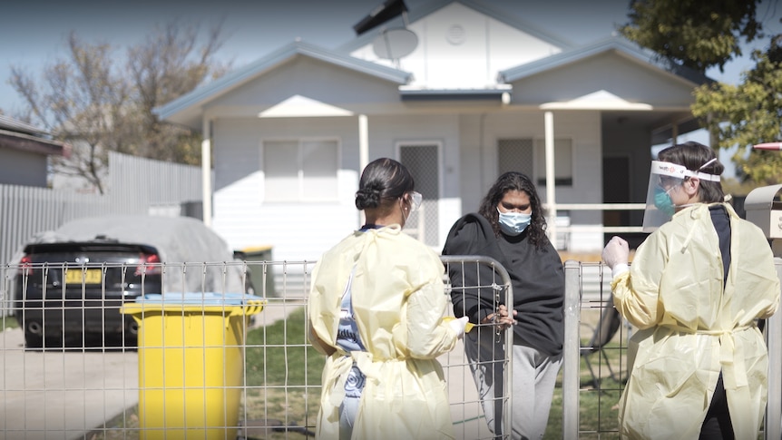 Two health workers in PPE talk to a woman over a fence in her front yard