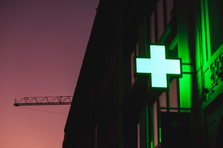 A green, glowing hospital cross sign outside a pharmacy 