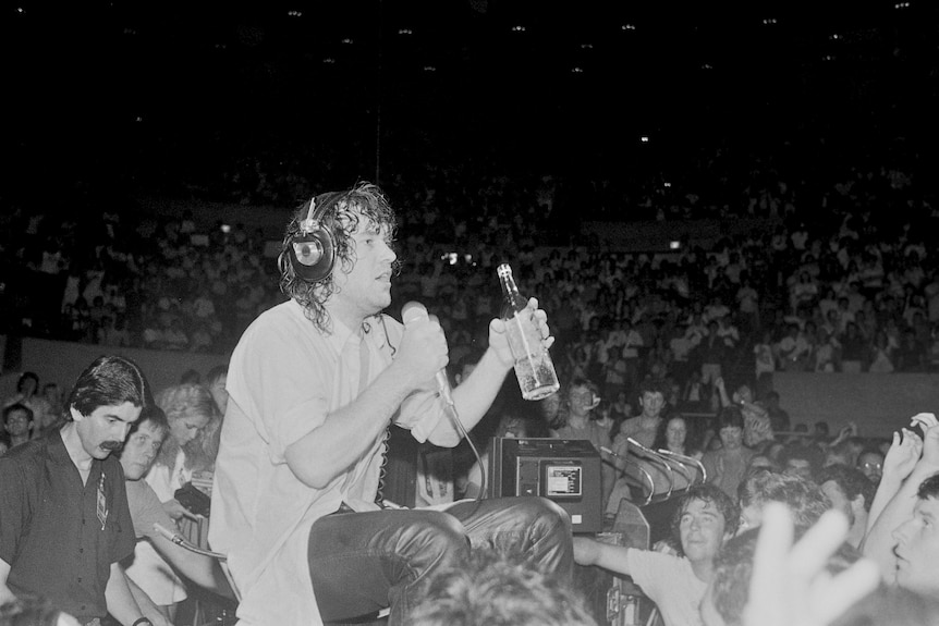 Black and white: Jimmy Barnes seated on stool on stage wearing headphone and holding a bottle of alcohol, surrounded by audience