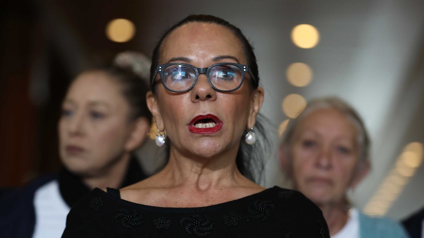 Linda Burney glances off camera while speaking to journalists. She is wearing dark red lipstick and black-framed glasses.