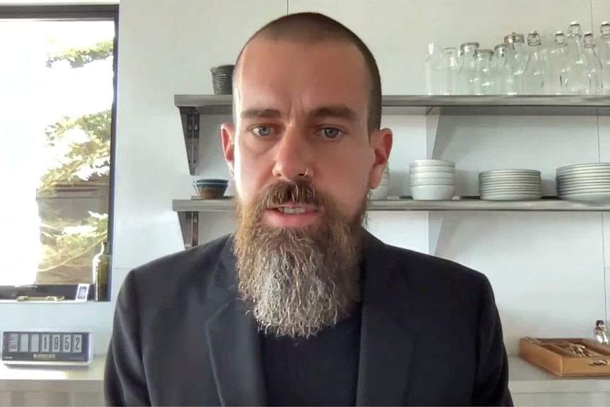 A man with a grey beard and wearing a dark suit stares at the camera as he sits