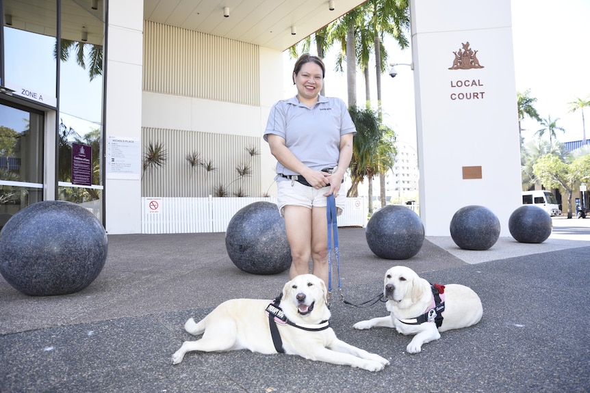 A woman stands outside the Darwin Local Court holding the leads of therapy dogs Roxy and Quinton lying on the ground. 