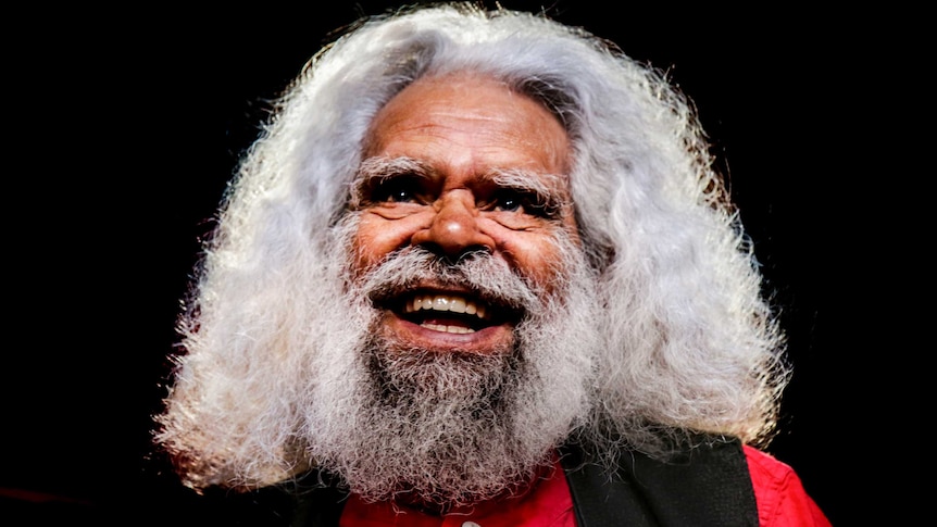 Jack Charles, wearing a red shirt and black vest, smiles before a dark black background.