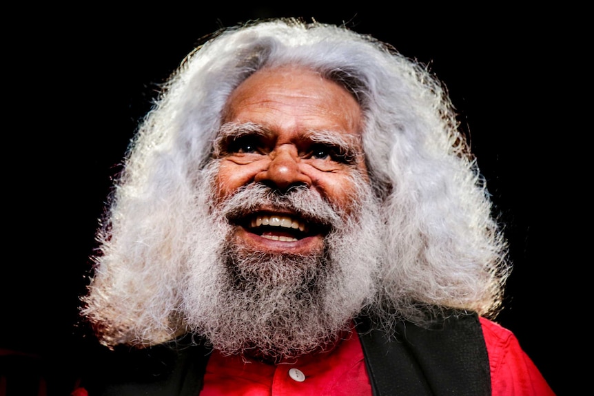 Jack Charles, wearing a red shirt and black vest, smiles before a dark black background.