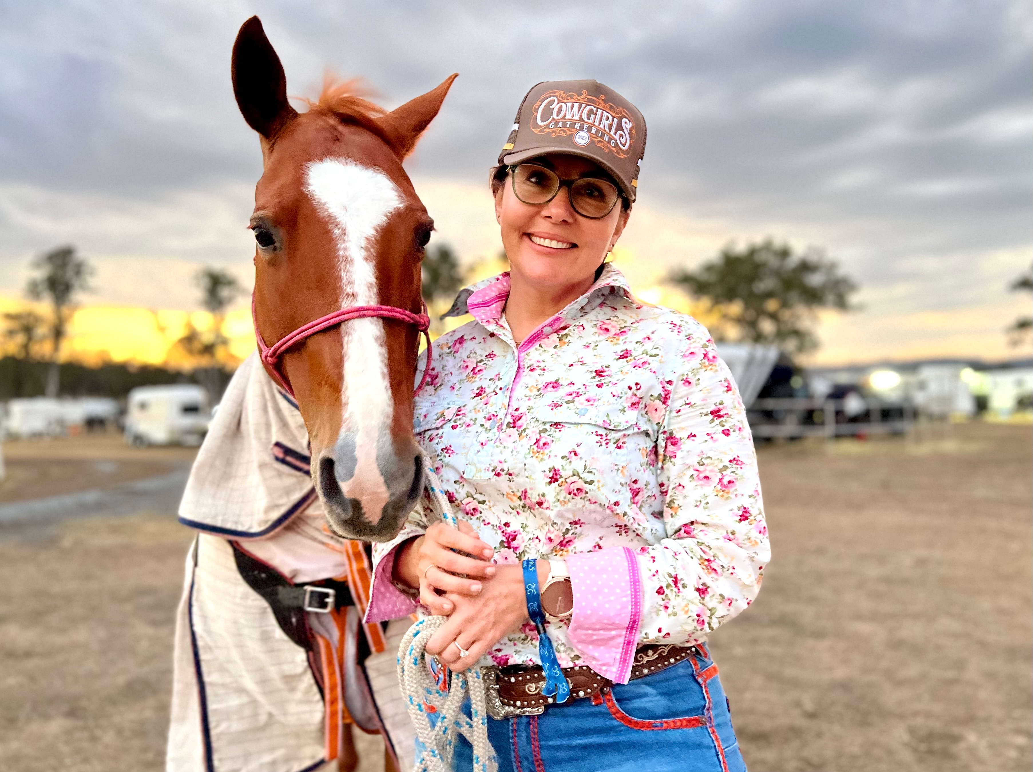 A woman in a floral shirt and cap poses next to a horse as the sun sets.