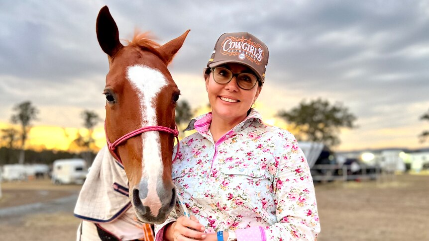 A woman in a floral shirt and cap poses next to a horse as the sun sets.