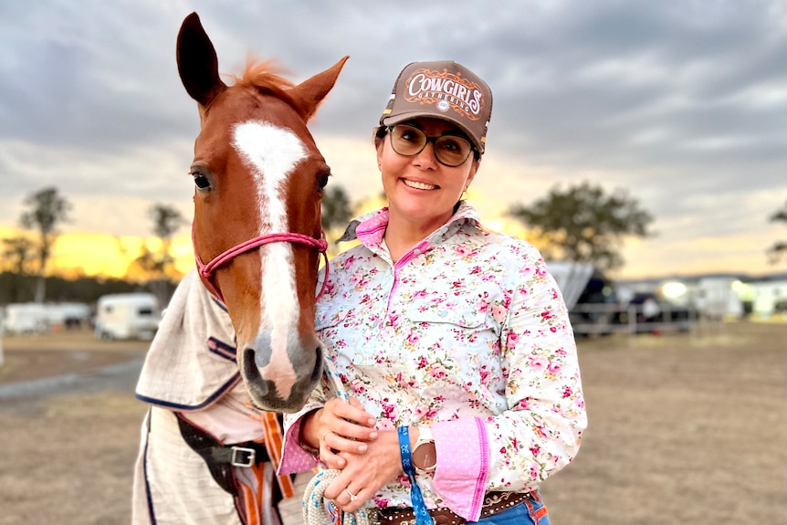 A woman in a floral shirt and cap poses next to a horse as the sun sets.