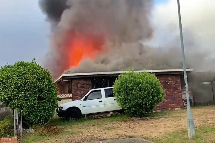 A single-storey home on fire with flames and smoke rising from the roof with a white car parked outside.