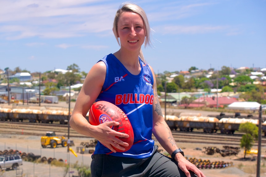 A blonde white woman wearing a blue singlet while holding an AFL football.