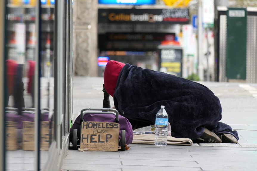 A person kneeling on a footpath under a blanket with a sign saying 'homeless please help'
