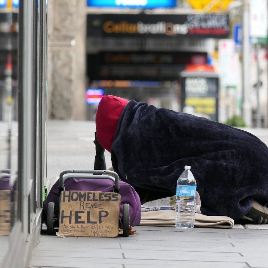 A person kneeling on a footpath under a blanket with a sign saying 'homeless please help'