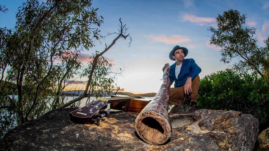 Lloyd Saunders kneels with a didgeridoo in his hand and a sunset background behind him.