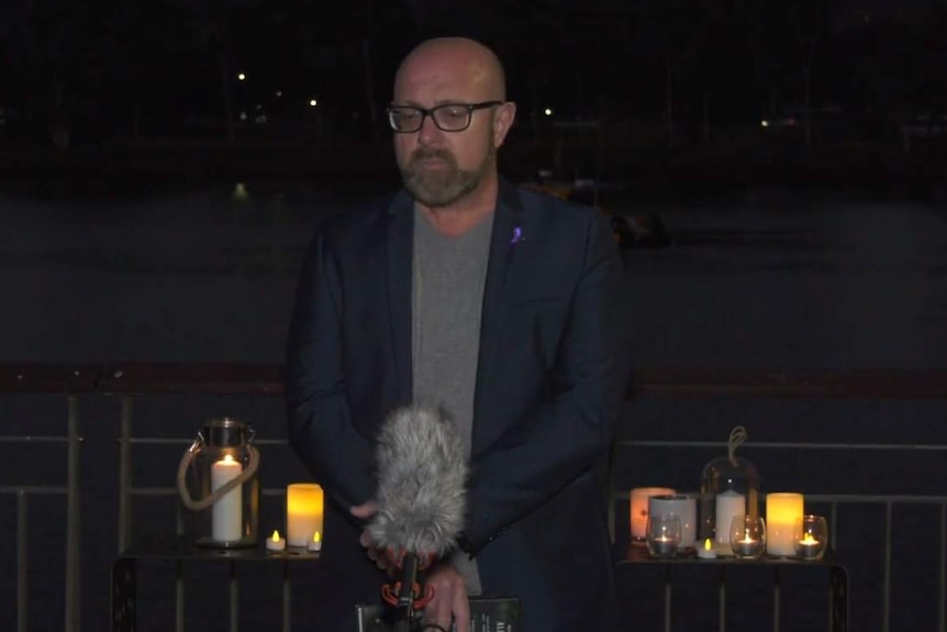 Bryan Cranston (Karen Gilliland's brother) stands with microphone, sombre look on face, candles and night sky behind.