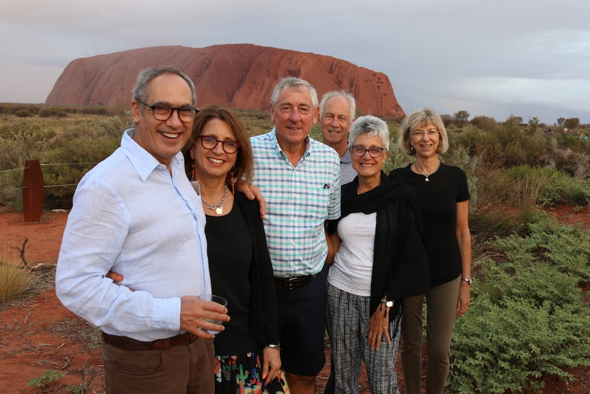A group of tourists pose for a photo, with Uluru in the distance.