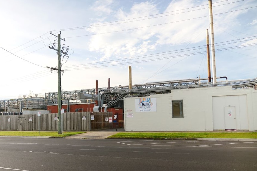A small white brick building surrounded by metal pipes and other factory equipment.