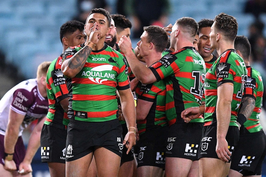 A South Sydney NRL player puts a finger to his mouth as he celebrates a try with his teammates in the background.