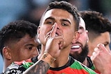 A South Sydney NRL player puts a finger to his mouth as he celebrates a try with his teammates in the background.