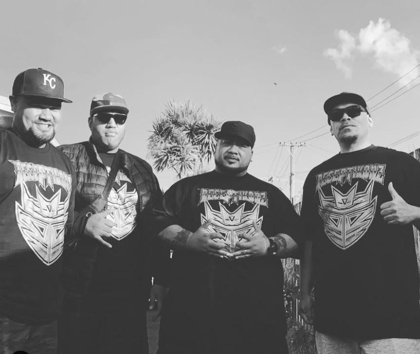 A black and white photo of four Polynesian men wearing black tee shirts standing outside with sky behind them. 