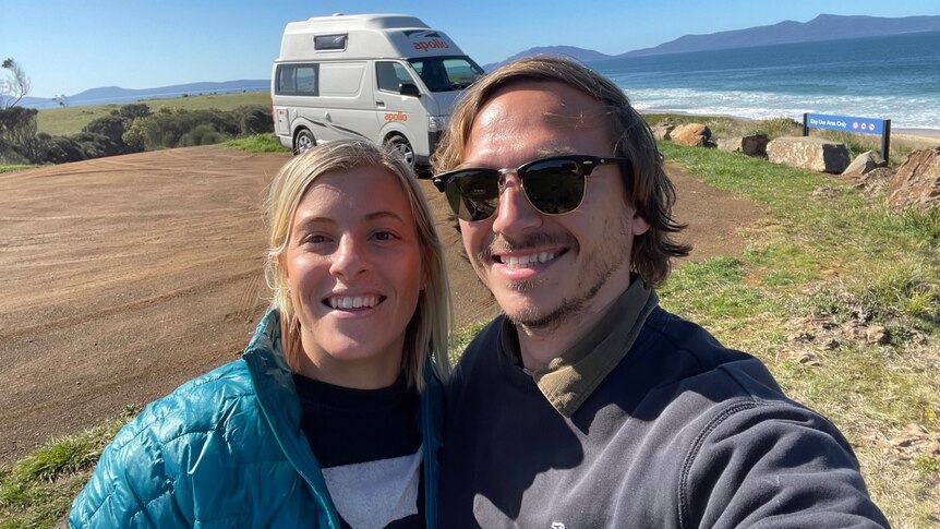 A blonde woman wearing a blue puffer jacket and a blonde man with black sunglasses taking a selfie at a beach