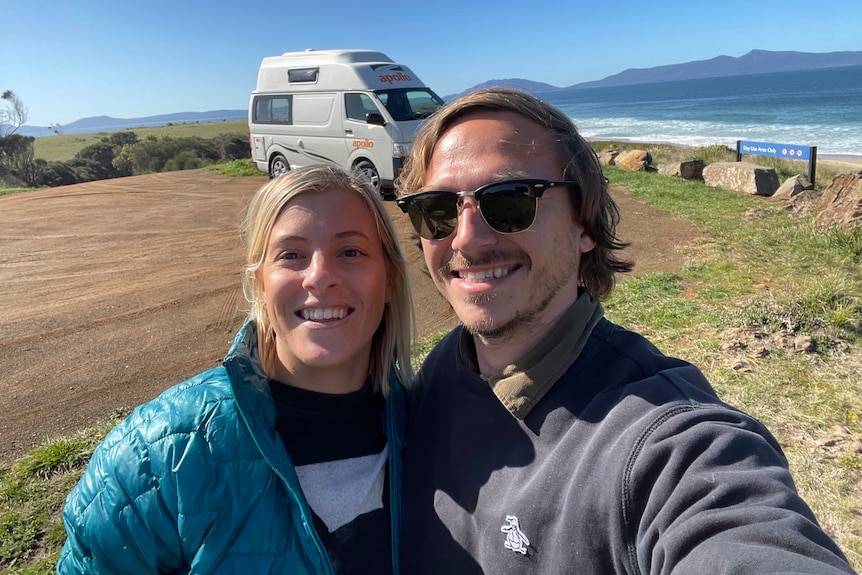 A blonde woman wearing a blue puffer jacket and a blonde man with black sunglasses taking a selfie at a beach