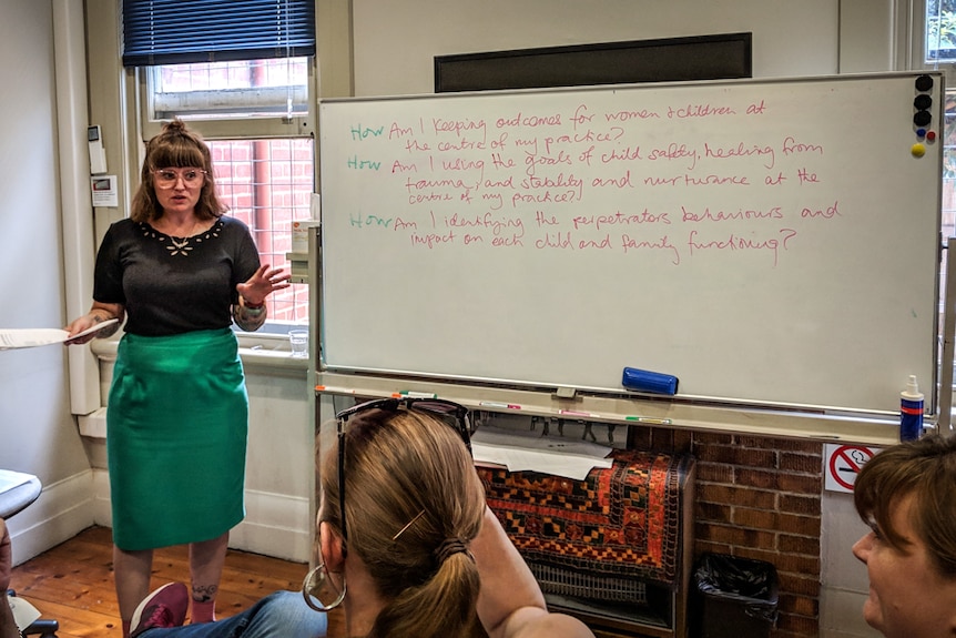 A woman stands alongside a whiteboard, covered with notes, delivering a talk to a group of seated onlookers.