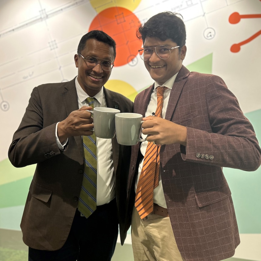 two dark-skinned men with spectacles in sports coats holding grey tea cups together
