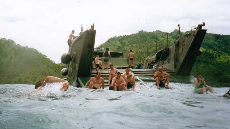 Soldiers sitting on and swimming in water around a military landing transport with islands in the background.