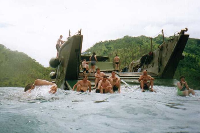 Soldiers sitting on and swimming in water around a military landing transport with islands in the background.