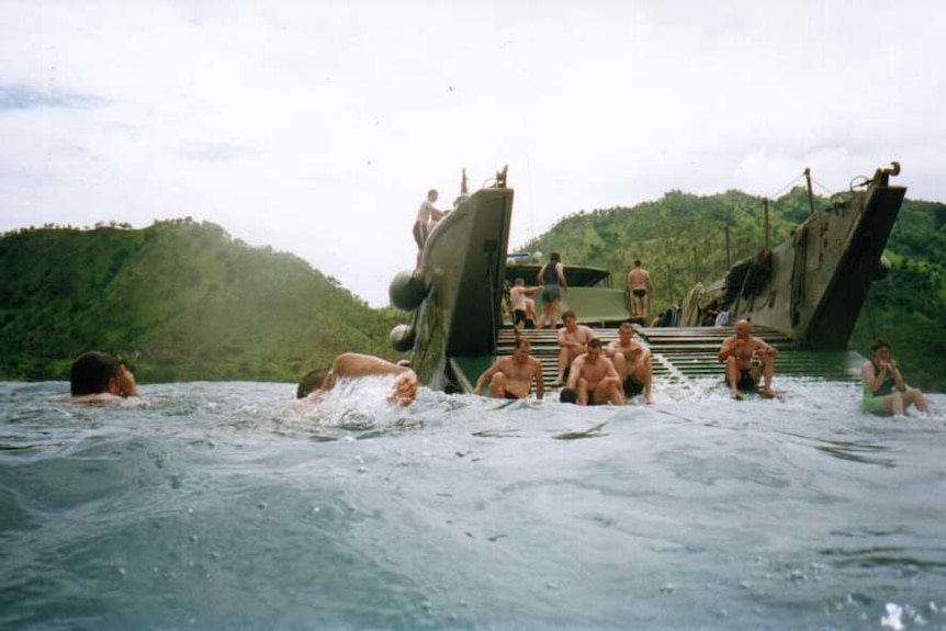 Soldiers sitting on and swimming in water around a military landing transport with islands in the background.
