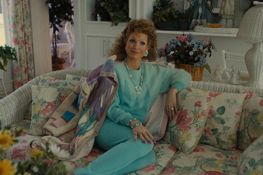 A blonde woman with an 80s perm, wearing a turquoise tracksuit, sits on a floral couch with an expectant expression