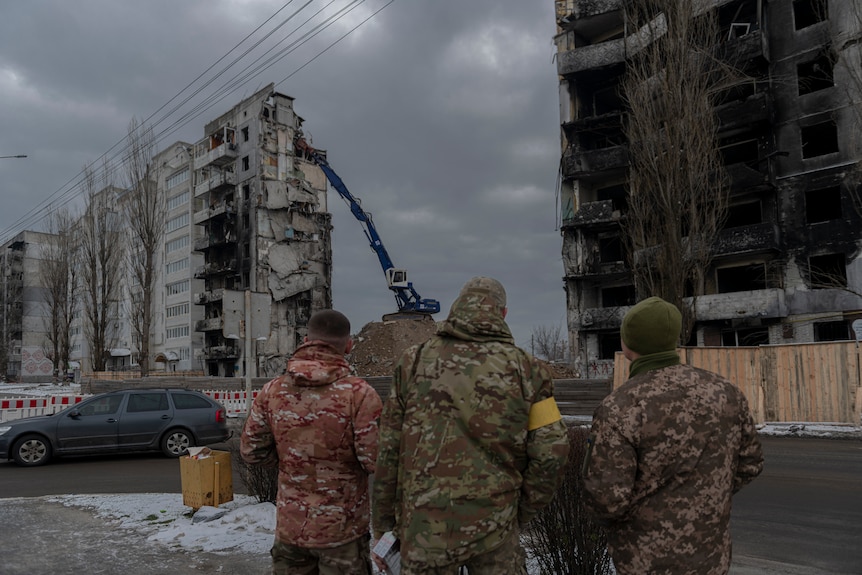 People in camouflage gear stand at a distance and watch a crane dismantle a building.