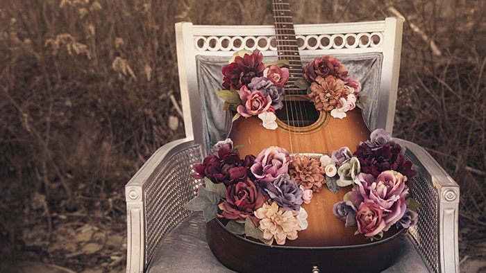 An acoustic guitar sits on a silver chair in a field, covered in pastel flowers.