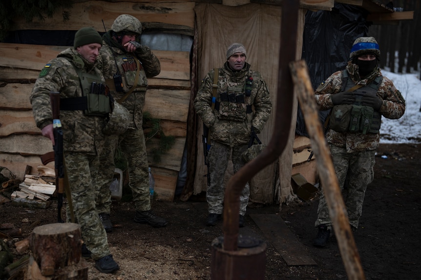 Four men in military uniforms lean against a wooden building. 