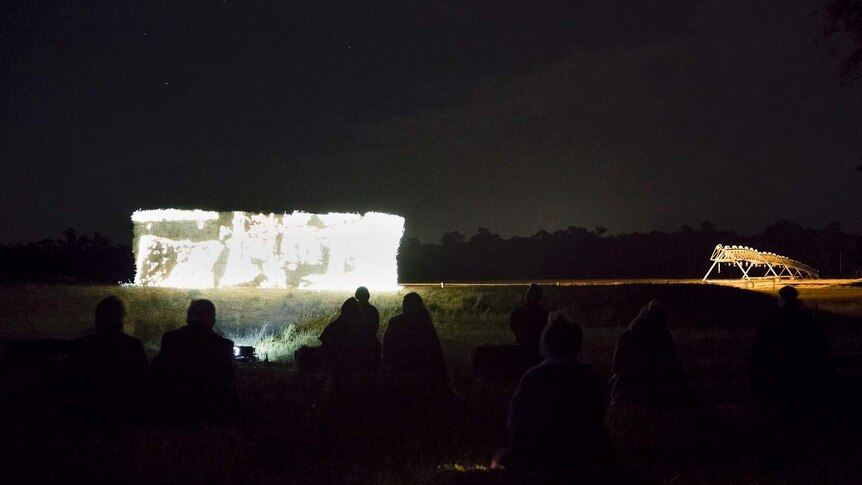 A group of people sitting on the grass looking at a visual artwork projected onto a stack of hay bales at night.