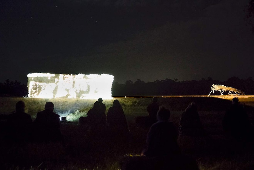 A group of people sitting on the grass looking at a visual artwork projected onto a stack of hay bales at night.