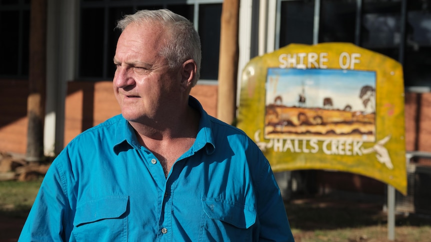 A man in a blue shirt looking away, Halls Creek sign in the background