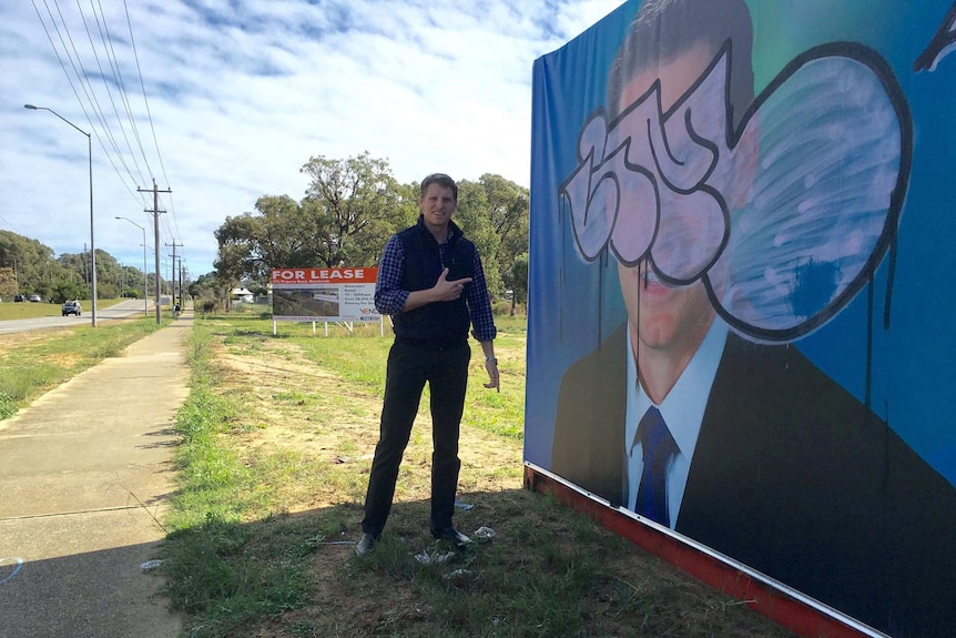 Andrew Hastie stands in front of a campaign banner showing his face with graffiti painted over it.