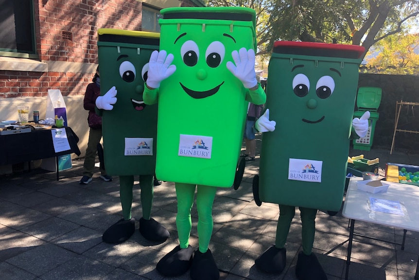 A green fogo bin mascot next to a recycling bin mascot and a landfill bin mascot outside  near trees