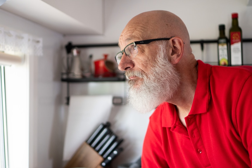 A man with a white beard looks out a window in a kitchen.