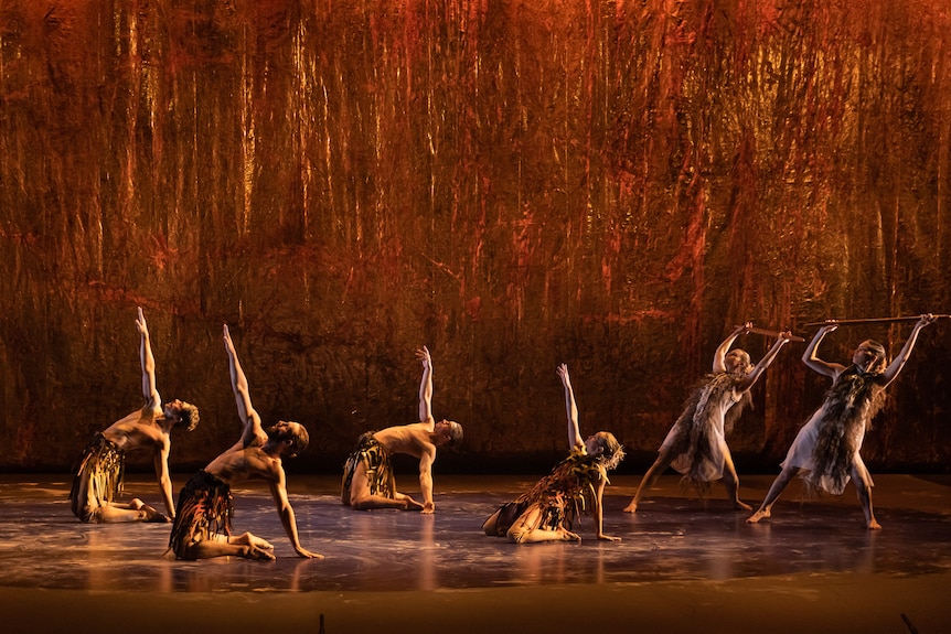 Six Indigenous dancers on stage, four on the ground and reaching up, two standing and holding staffs, bathed in a bronze light