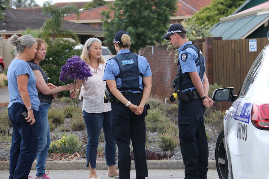 Three women holding a bunch of flowers talk to two police officers outside a house