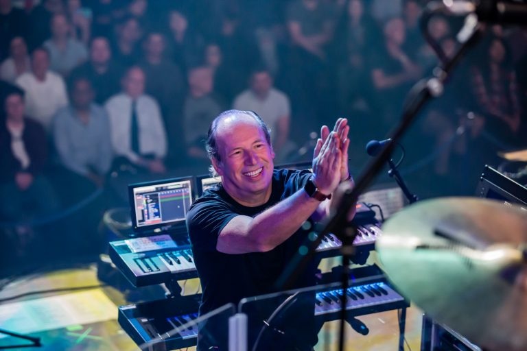 A man standing in front of keyboards, smiles and claps as an audience sits behind him.