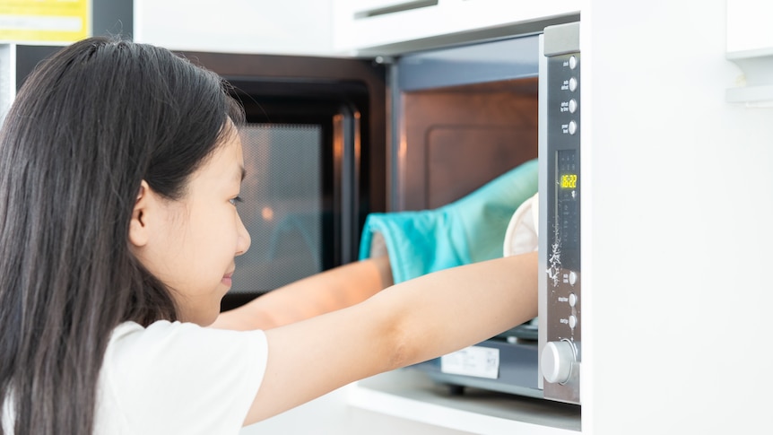 Young dark haired woman pulls something out of microwave wearing gloves, in a story about surprising microwave uses.