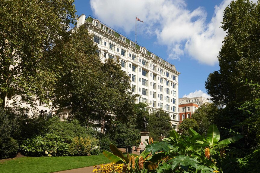 Exterior of the Savoy Hotel in London on a bright sunny day. The hotel is grand in size and surrounded by lush green gardens.