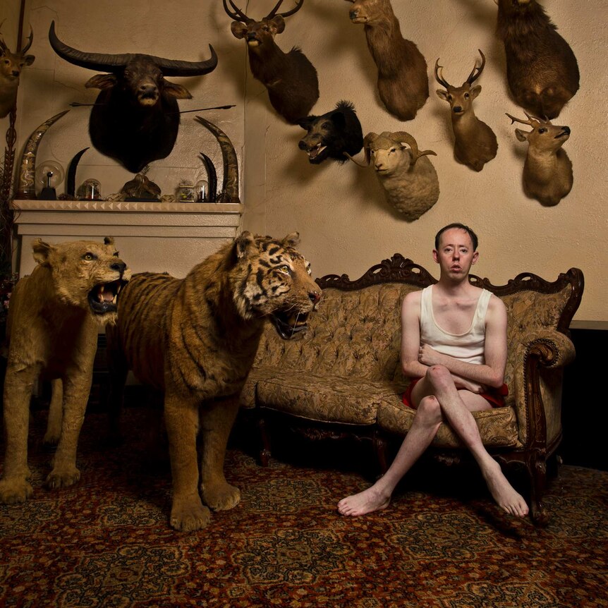 A man in a singlet sits on a couch in a room full of taxidermied animals.