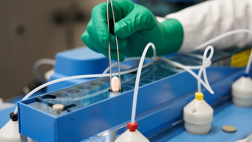 A gloved hand holds tweezers placing a pill in a vial on a scientific benchtop