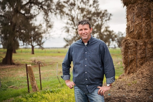 A man in a dark, long-sleeved shirt stands on a country property in front of a tree.