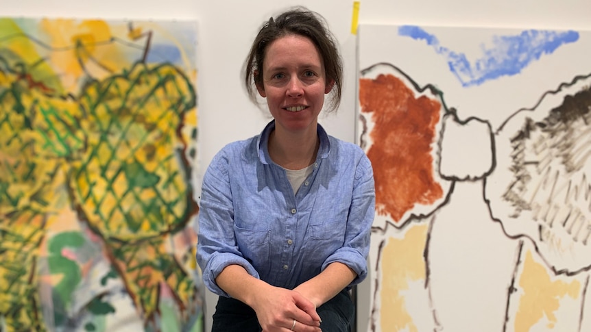 Artist Kate Smith sits in front of two painted canvases in an art space